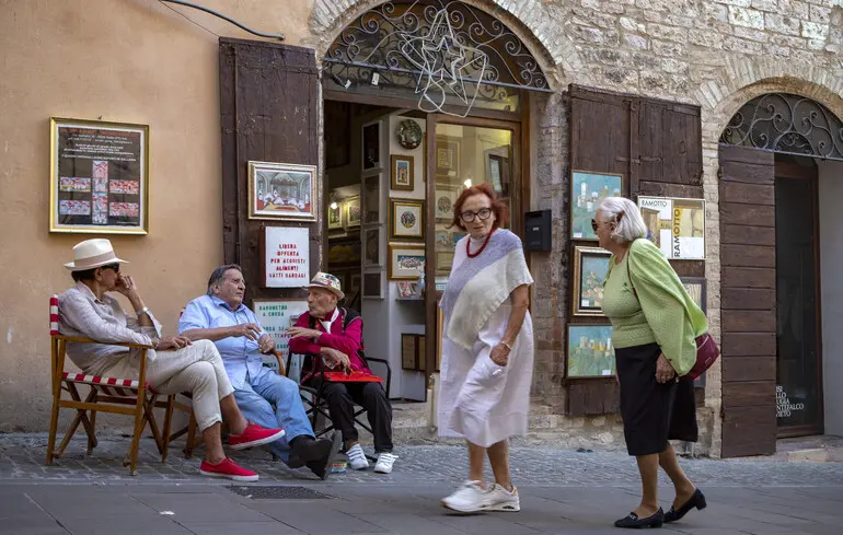 Літні італійські чоловіки і жінки розмовляють біля художньої галереї в стародавньому місті Спелло, регіон Умбрія, Італія / 17 вересня 2025 року